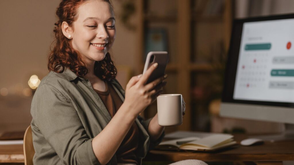 How to advertise on ChatGPT Ads Manager: professional smiling while using a phone in front of a computer, holding a coffee