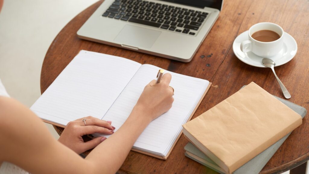 Como escrever artigo para blog: pessoa escreve num caderno ao lado do notebook e de uma xícara de café